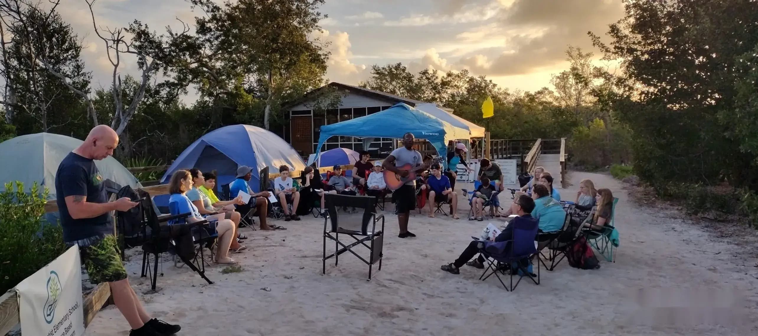 Students and teachers gather in a circle for evening worship at a beach campsite
